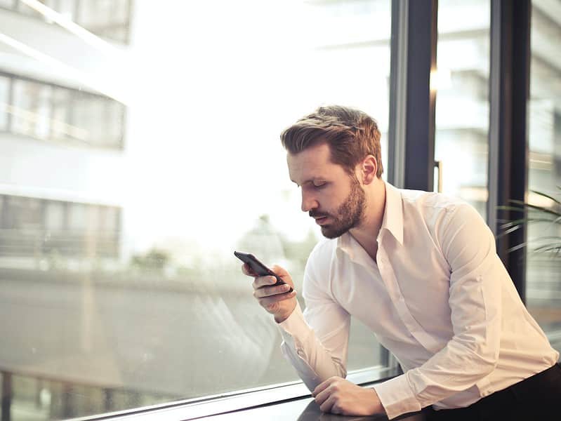 photo of man in white dress shirt holding phone near window